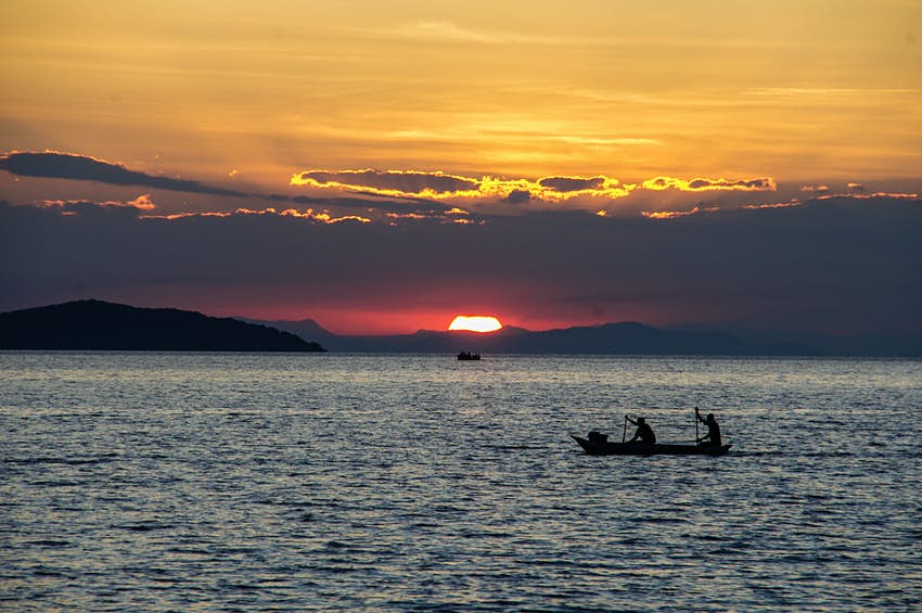 Dos pescadores en una canoa reman en un lago mientras se pone el sol, cubriendo el cielo de naranja y violeta