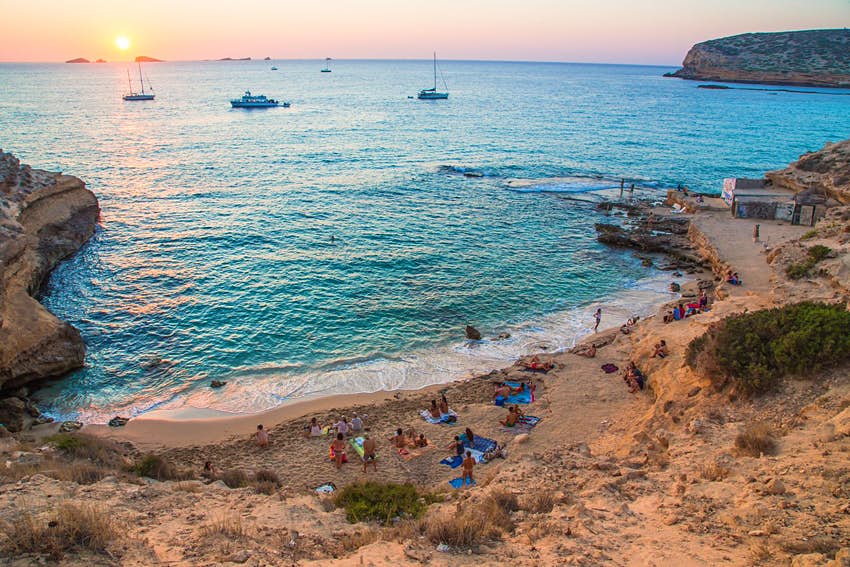 La gente se alinea en la arena de la playa de Cala Compte, Ibiza.  Varias personas se sientan en toallas de colores mientras una pareja nada en el mar.  El sol se pone en el fondo.