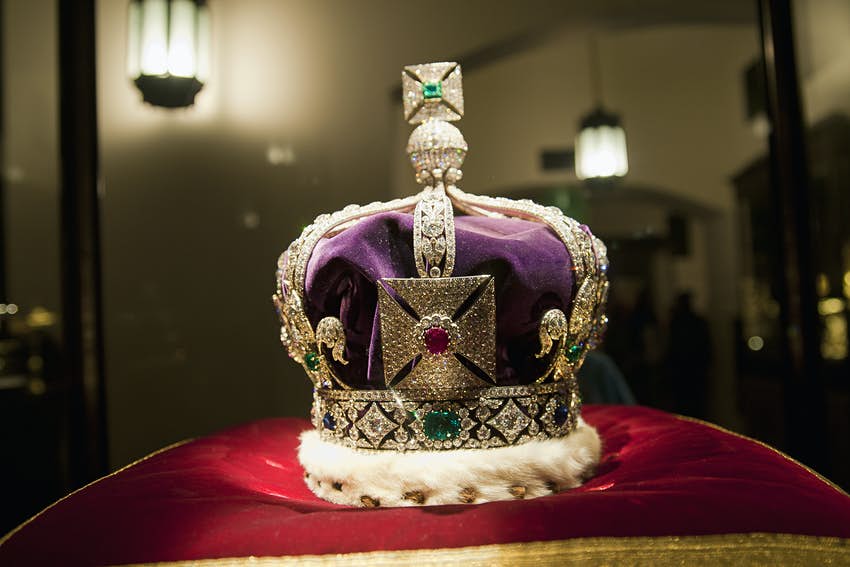 Sobre una almohada roja se encuentra una corona cubierta con joyas grandes y pequeñas. 