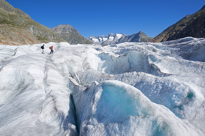Dos turistas caminan por el glaciar Alech, un paisaje accidentado y helado.  Sobre ellos el cielo es azul brillante.