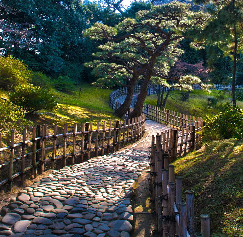 Una cerca de madera rodea un camino de piedra que atraviesa un pueblo tranquilo en Ritsurin Cohen, en la ciudad japonesa de Takamatsu.  Están todos los árboles dispersos en las colinas cubiertas de hierba.  