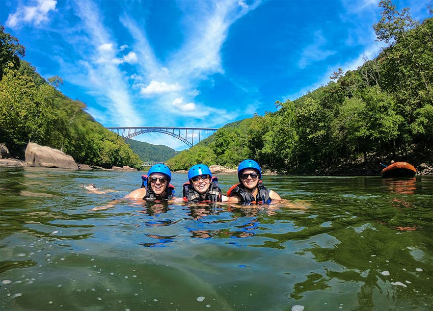 Tres mujeres nadan en un río bajo un cielo azul en un desfiladero bordeado de árboles.  Un puente de arco cruza el río en el fondo