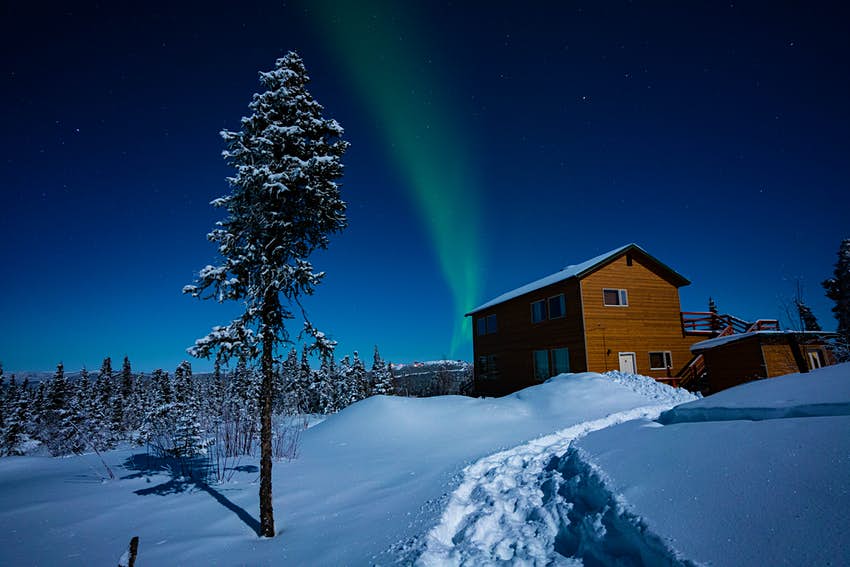 Una escena nevada de una cabaña de madera rodeada de bosque, mientras un destello de rayas verdes perfila el cielo arriba.