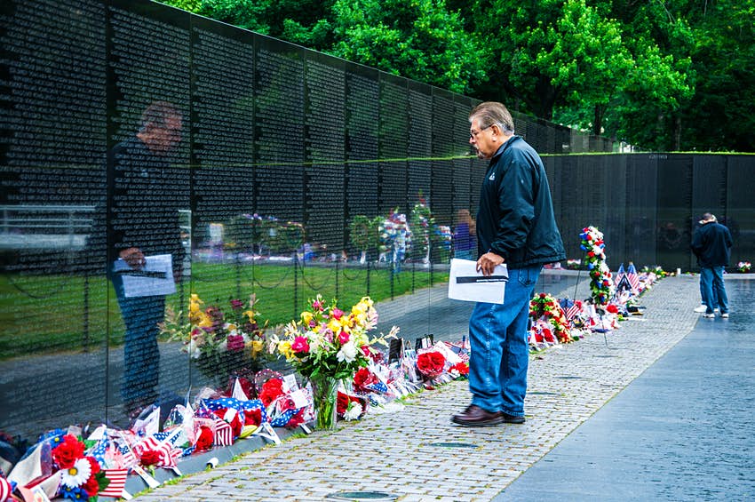 Un hombre se para frente al monumento.  Hay flores por todo el frente de la pared.