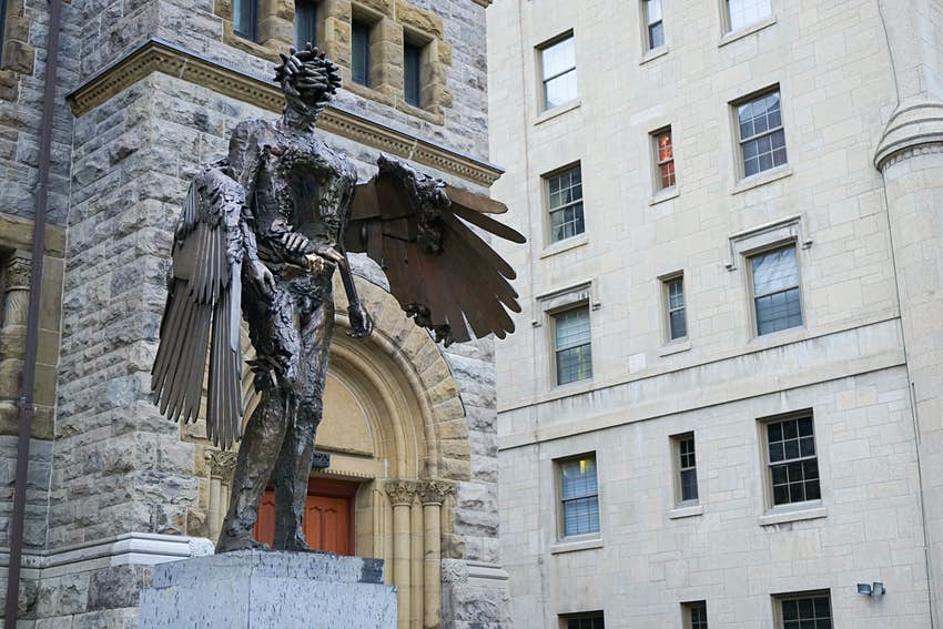 Una gran escultura de metal de la figura humana alada se encuentra sobre un pedestal fuera del edificio.
